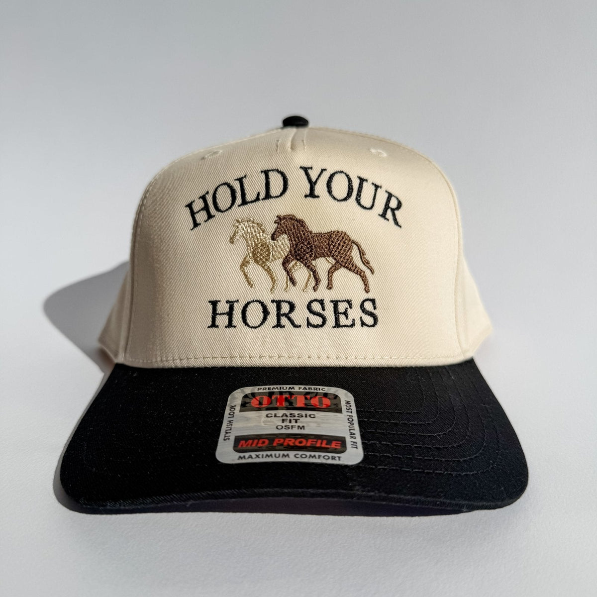 Front view trucker hat: beige panel, black brim, “HOLD YOUR HORSES” and horse emblem