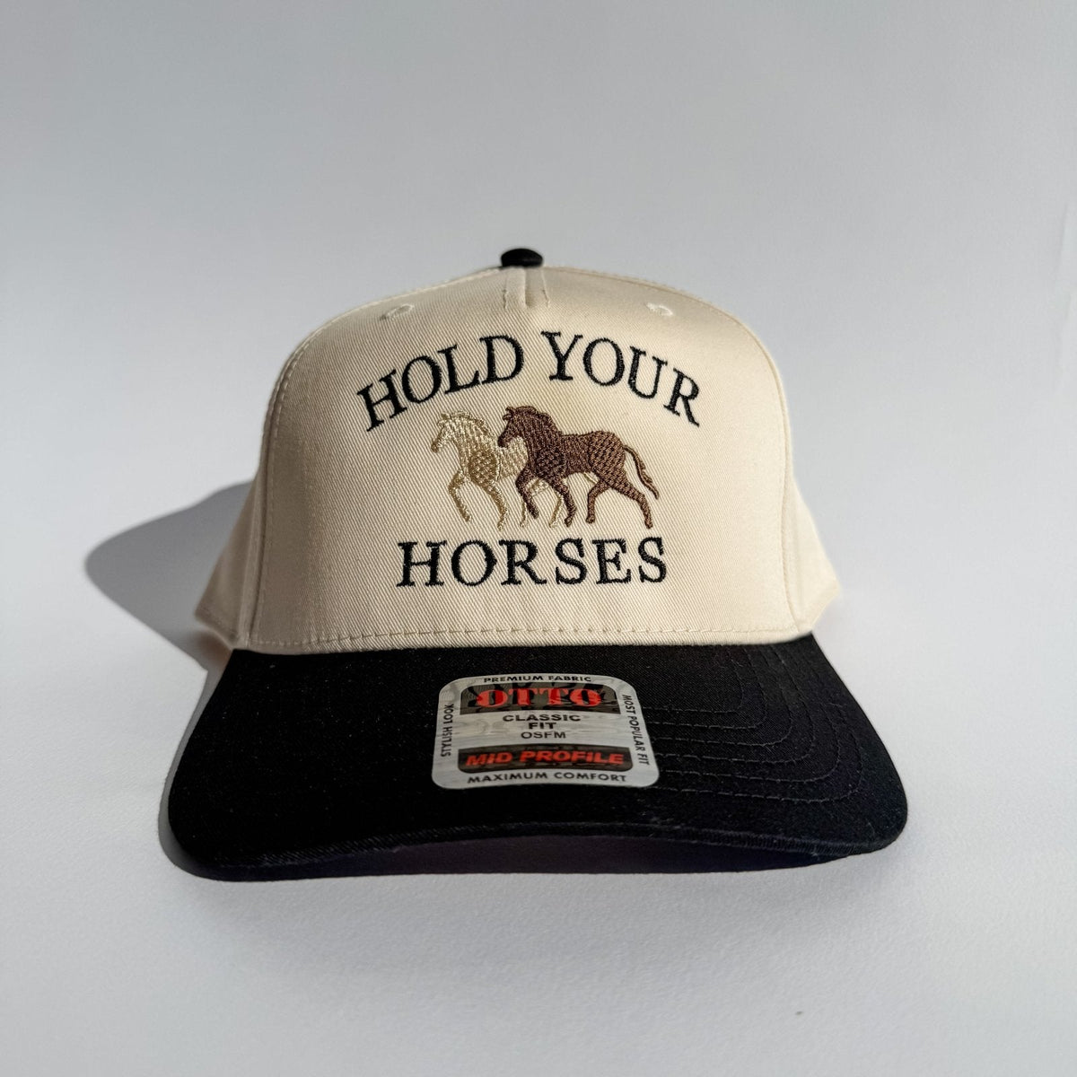 Beige and black trucker hat with “HOLD YOUR HORSES” lettering and horse graphic