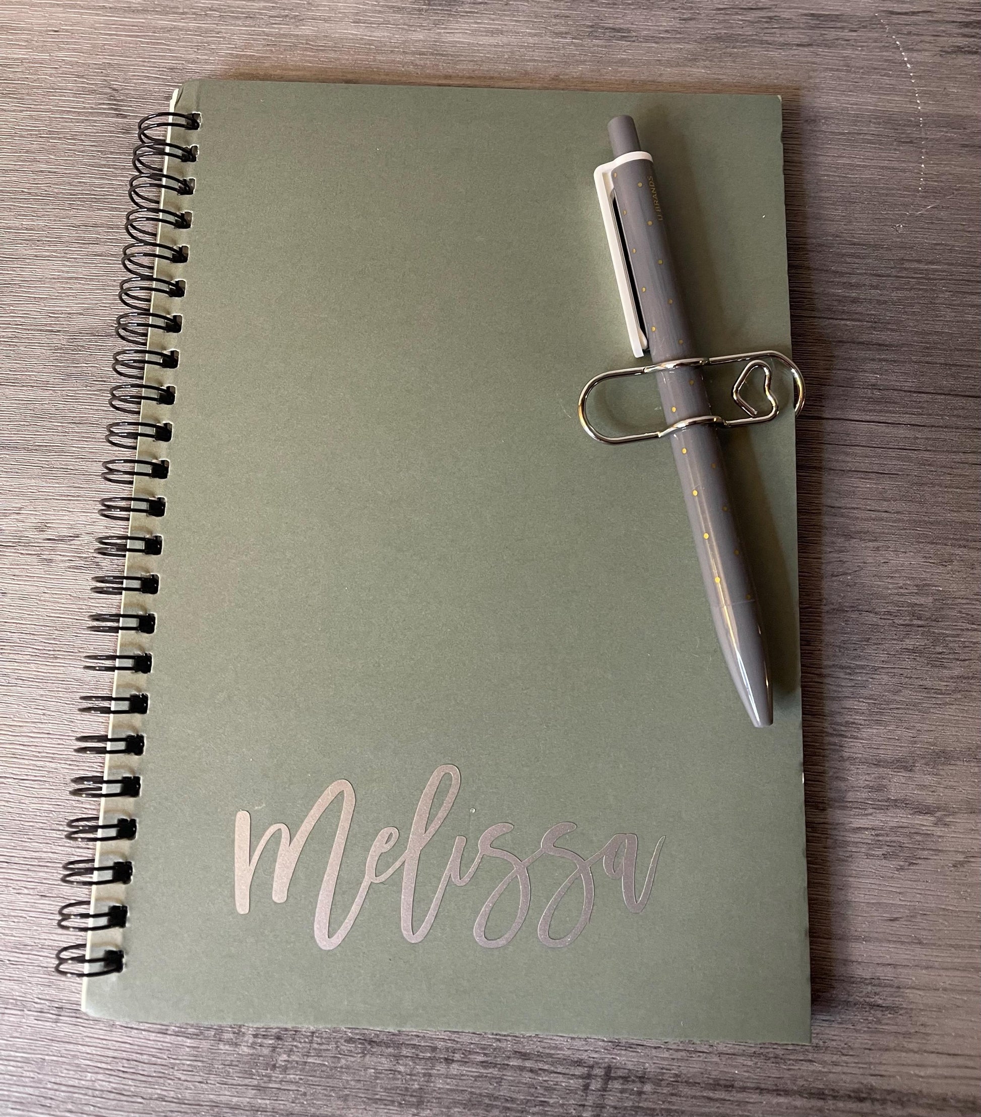 Olive spiral notebook with pen clip and script name 'Melissa'.