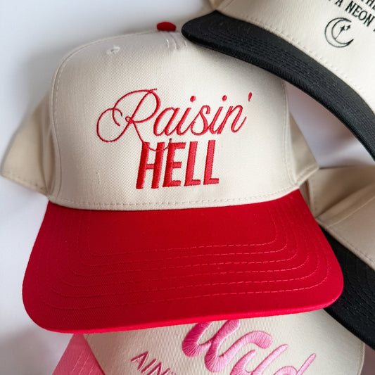 Cream and red trucker hat with embroidered "Raisin' Hell" and black mesh back